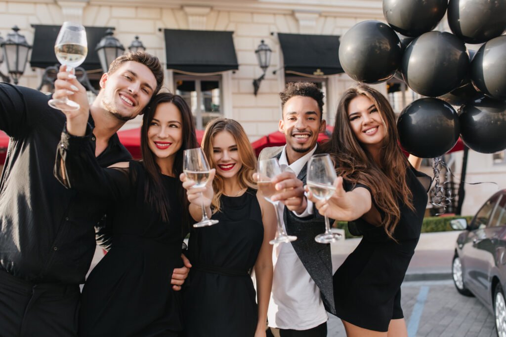 graceful girl with long light brown hair holding black helium balloons in front of building and laughing. cute blonde woman raising glass of champagne while posing with african friend..